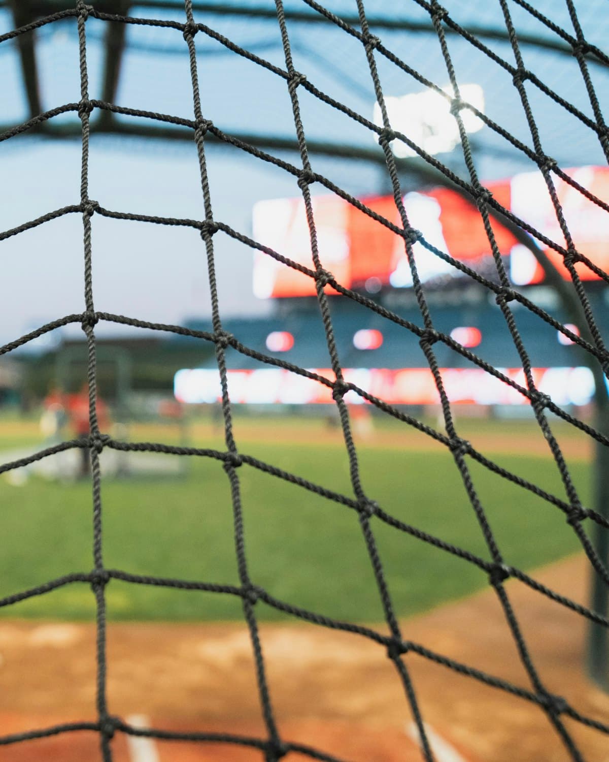 A batting cage at dusk, the net in the foreground and the field lights beyond.